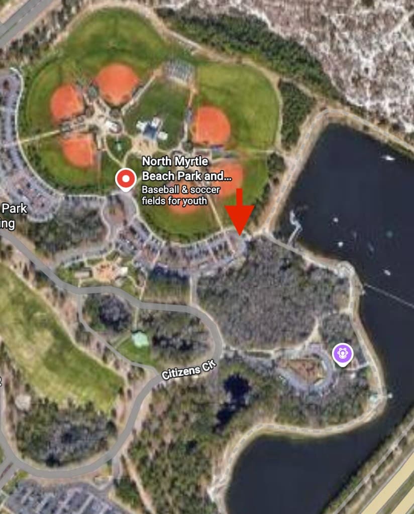 Satellite map of North Myrtle Beach Park and Sports Complex showing the recommended venue meetup area marked with a red arrow at the southeastern edge of the curved parking lot by the lake